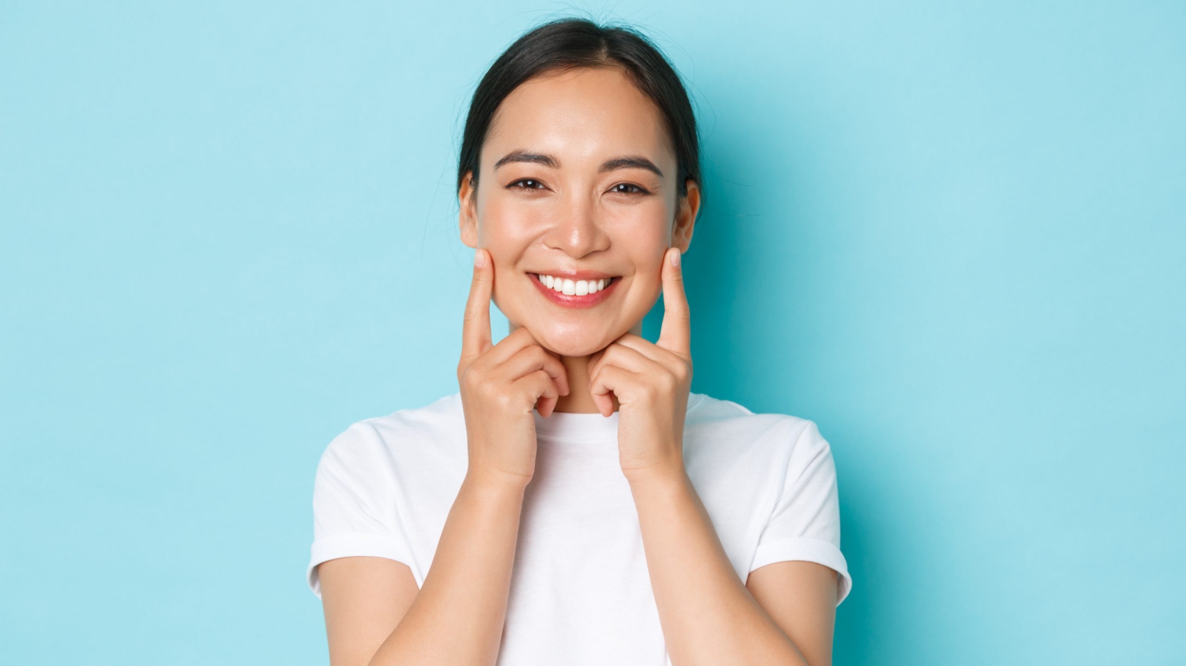 Close-up of beautiful asian girl looking pleased, smiling white teeth, touching cheeks, recommend skincare product, cosmetics for facial skin, standing light blue background.