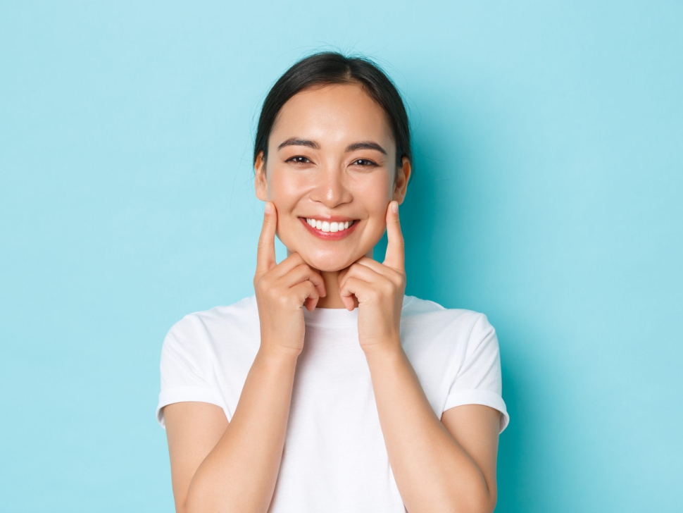 Close-up of beautiful asian girl looking pleased, smiling white teeth, touching cheeks, recommend skincare product, cosmetics for facial skin, standing light blue background.